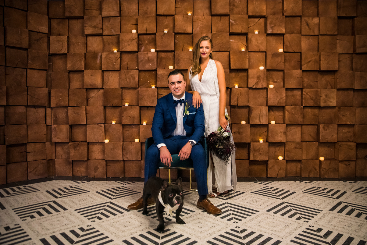 A bride and groom pose with their dog in front of a wooden wall at The Oaks at Plum Creek.