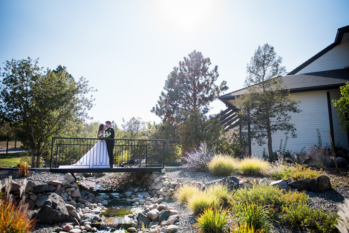 A bride and groom on a bridge in front of their Denver wedding venue, The Oaks at Plum Creek.