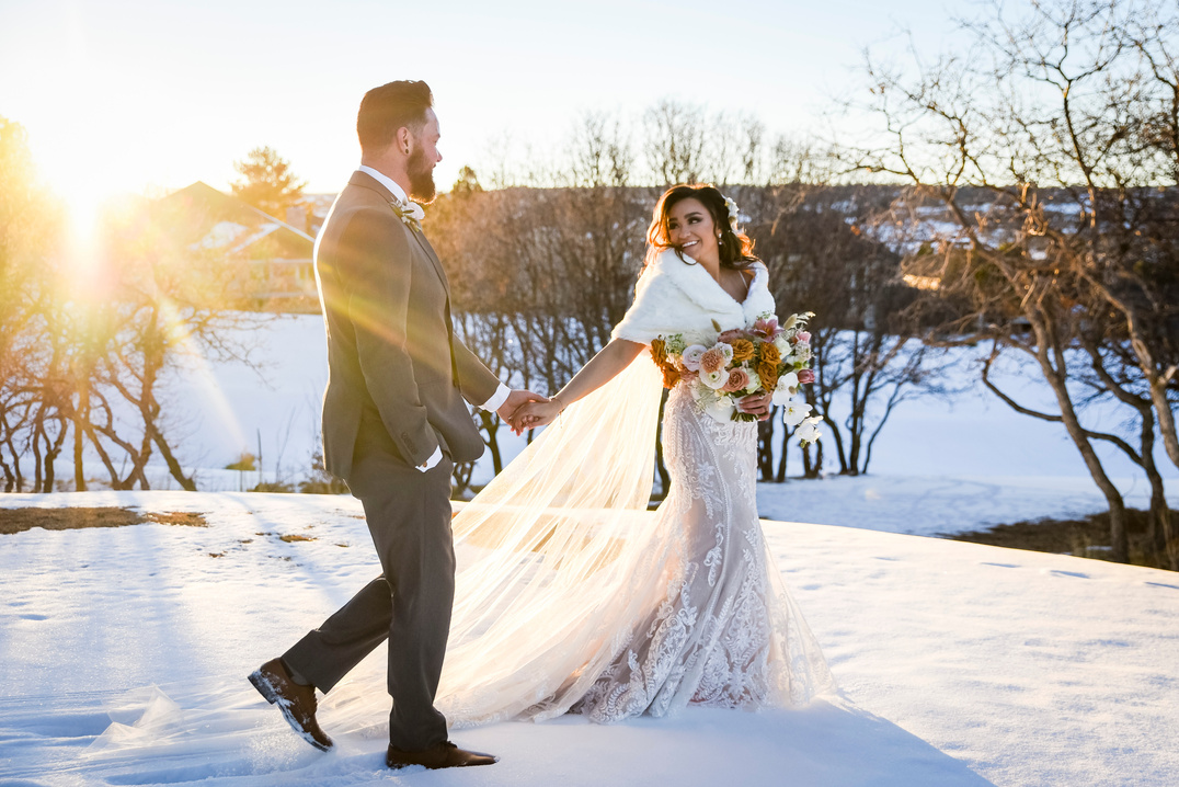 A bride leads her groom walking in the snow at sunset at The Oaks at Plum Creek in Castle Rock, Colorado.