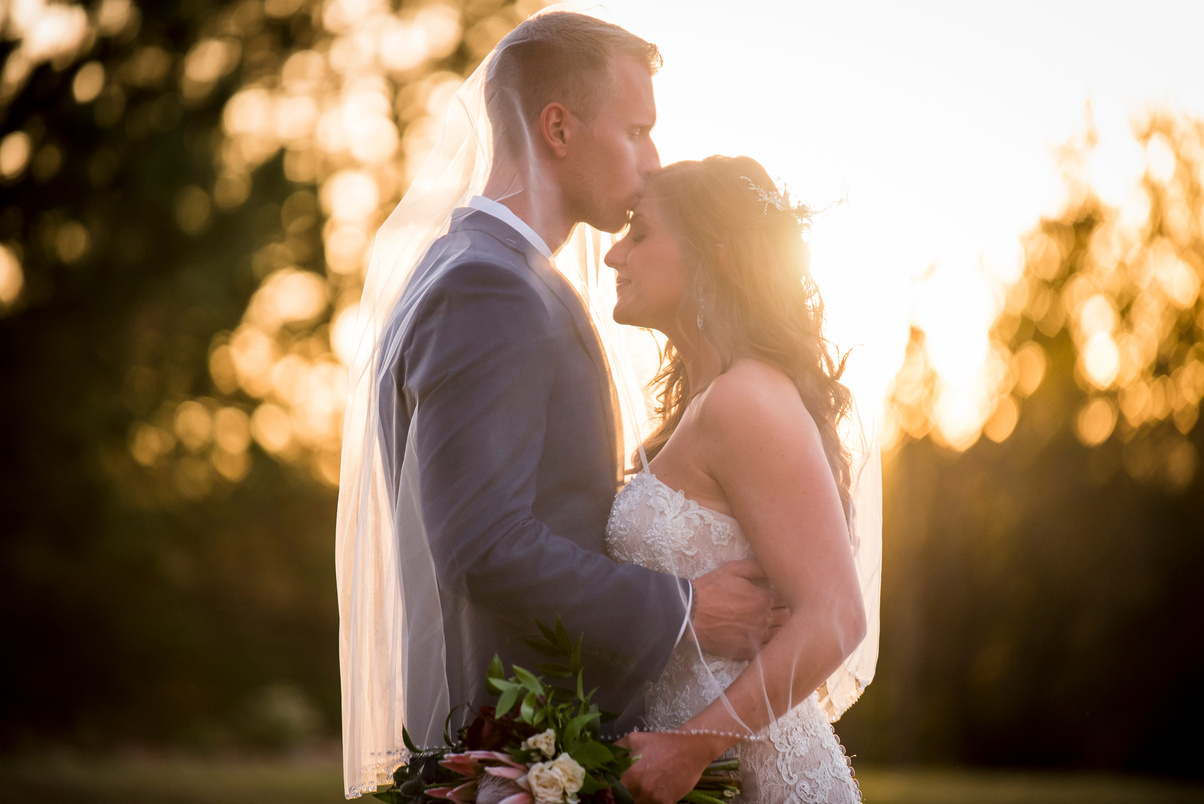 A groom kisses his bride on the forehead at sunset at The Oaks at Plum Creek.